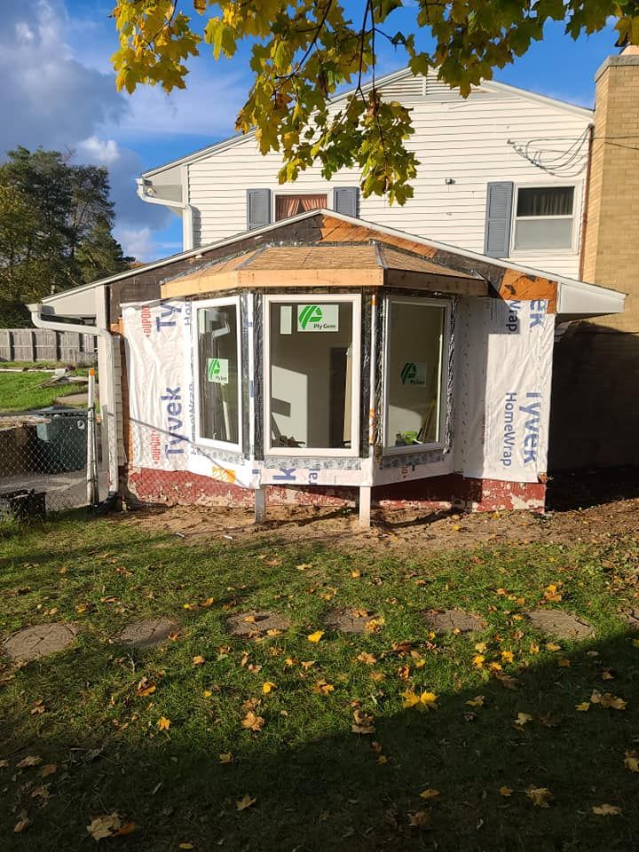 A partially constructed sunroom with a bay window attached to a two-story white house. Blue Tyvek wrap covers the frame, with a grassy yard in front.
