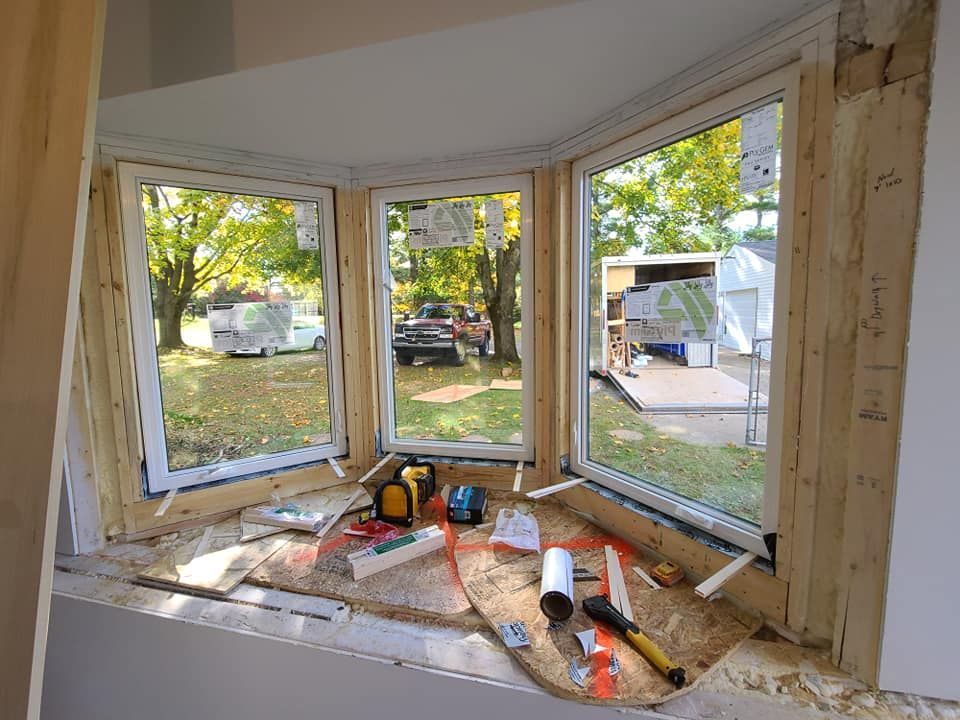 Bay window installation in progress, showing three white-framed windows with tools and materials on the windowsill. A view of the outdoors is visible.