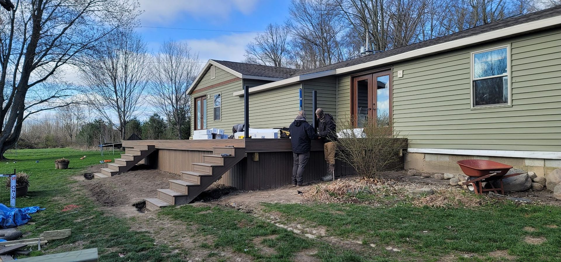 Two people near a deck of a house with green siding. A wheelbarrow sits near the house.