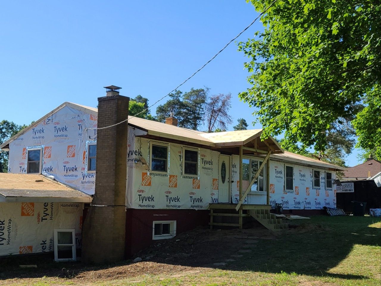 House under construction with Tyvek wrap visible. A brick chimney and wooden porch are present, set against a clear blue sky.