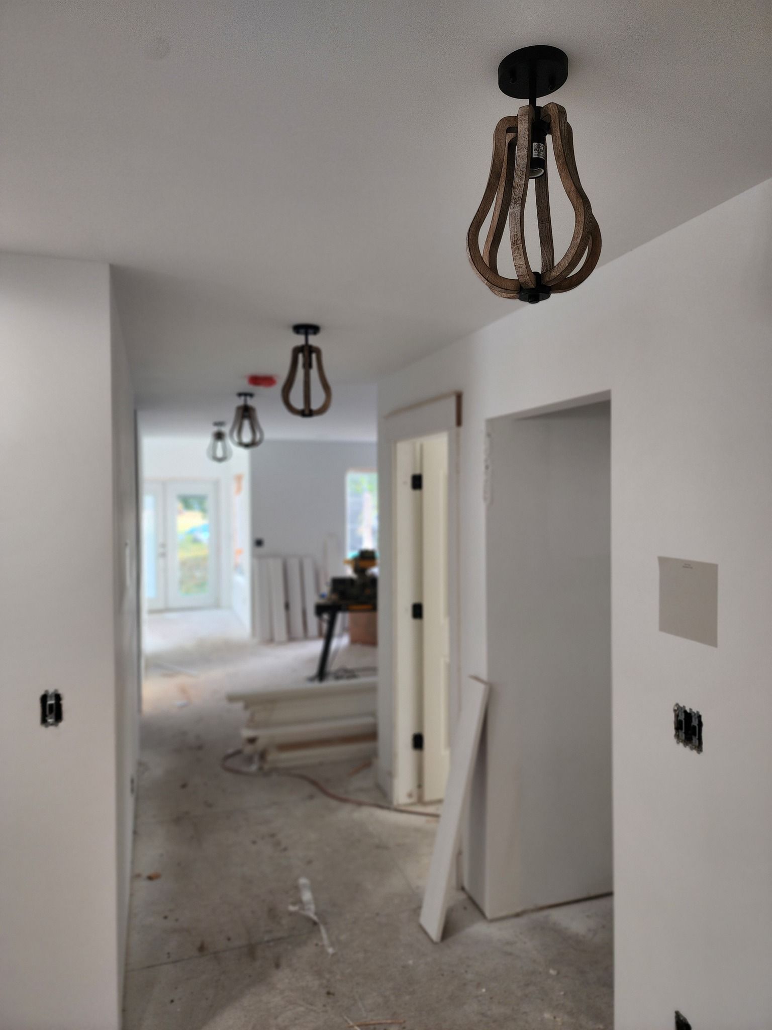 Hallway with three rustic pendant lights installed, leading to a room with a doorway and exterior view. Walls are painted white.