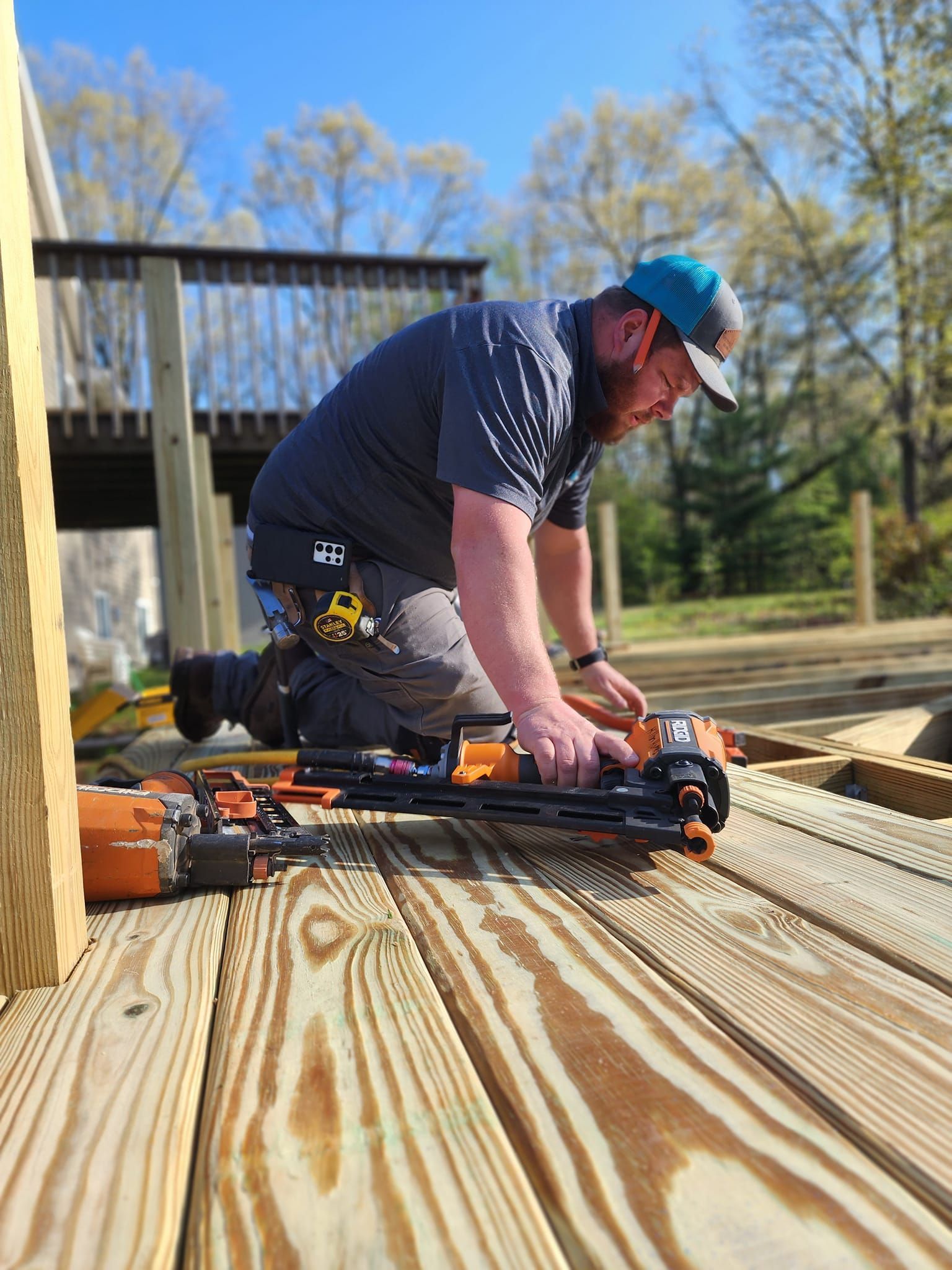 A construction worker kneels on a partially built wooden deck, using a nail gun. He wears a blue hat, gray shirt, and work pants.
