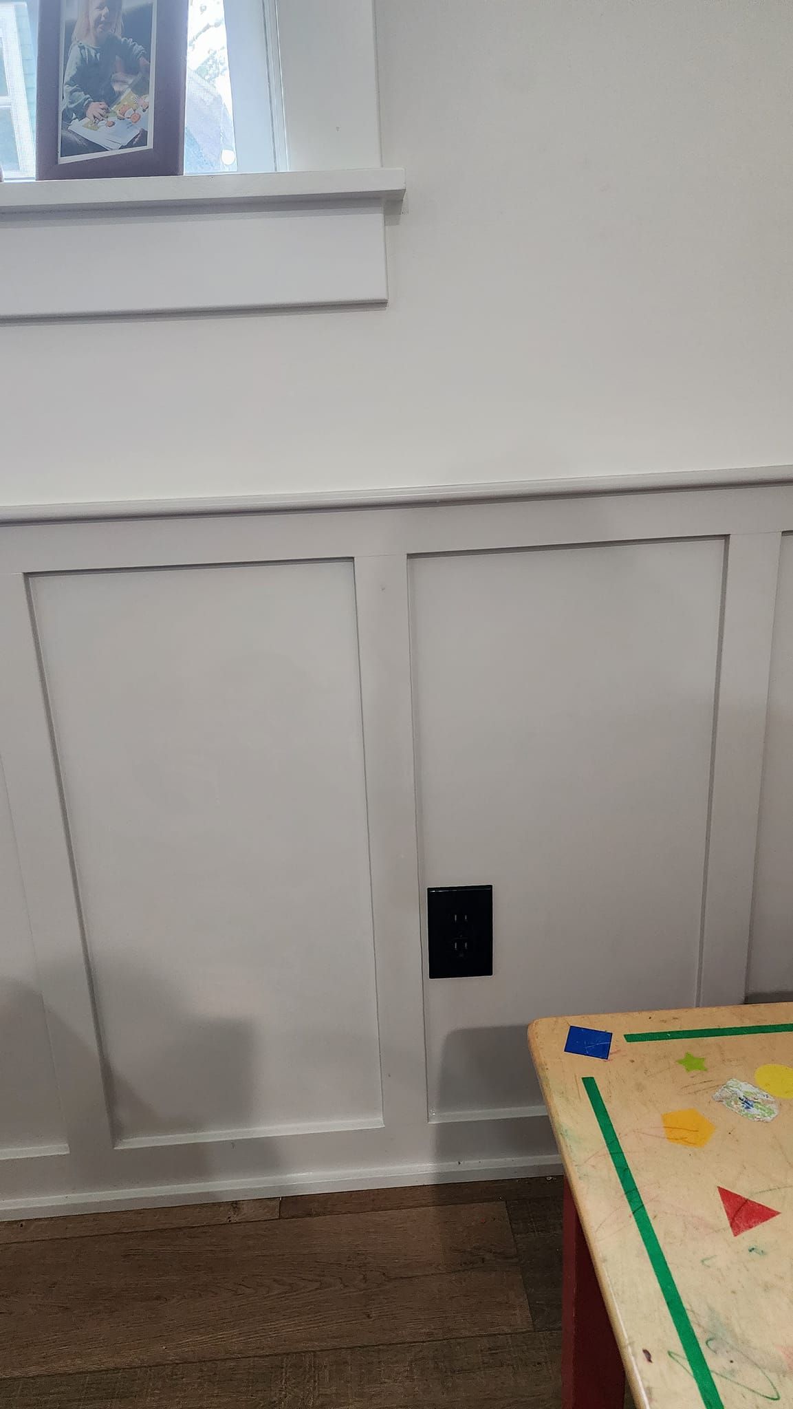 White paneled wall with an electrical outlet. A wooden play table sits in the foreground.