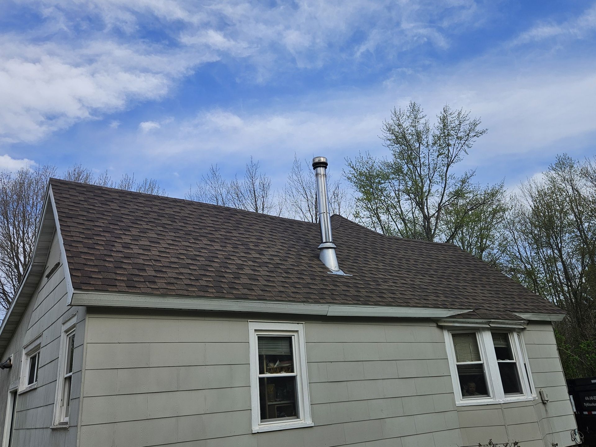 A house with a brown shingled roof and a metal chimney against a cloudy blue sky.