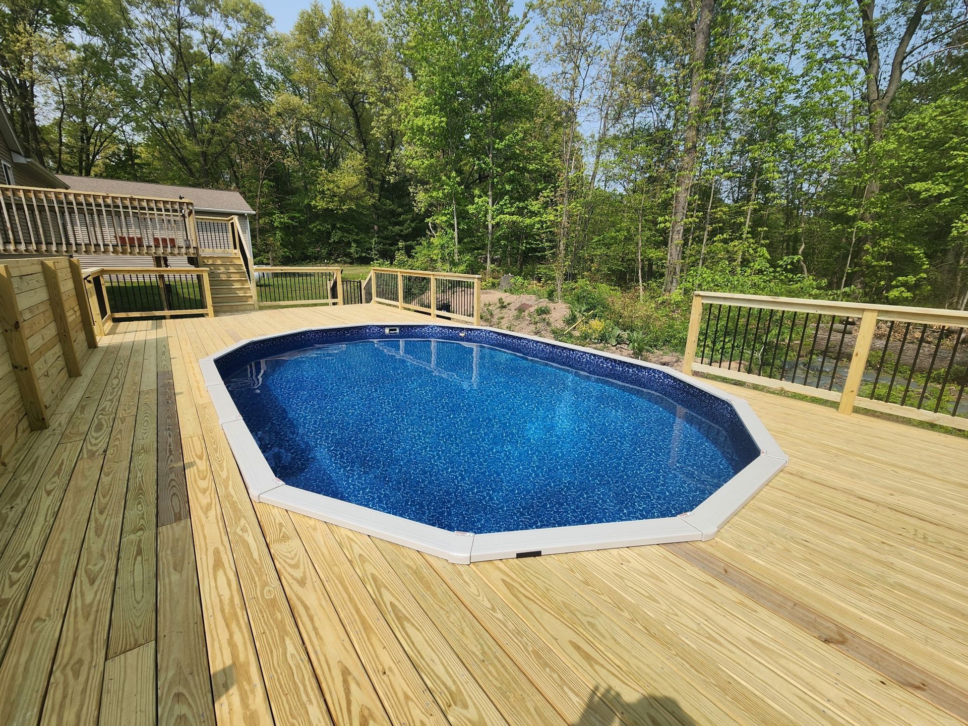 Oval swimming pool surrounded by a wooden deck and fencing. Green trees surround the pool in the background.
