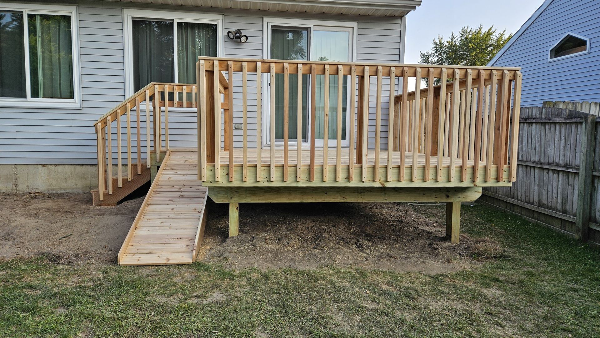 Newly built wooden deck with a ramp and railing, attached to a light gray house. A backyard setting with grass and a fence.