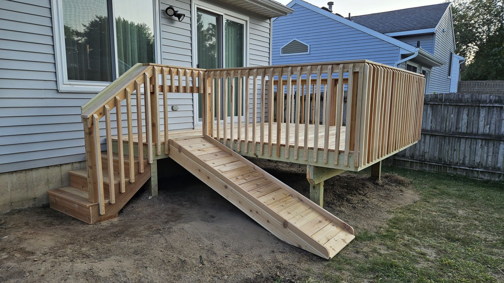 Wooden deck with stairs and a ramp leading to a grassy yard. A house with siding is in the background, and a wooden fence is to the right.