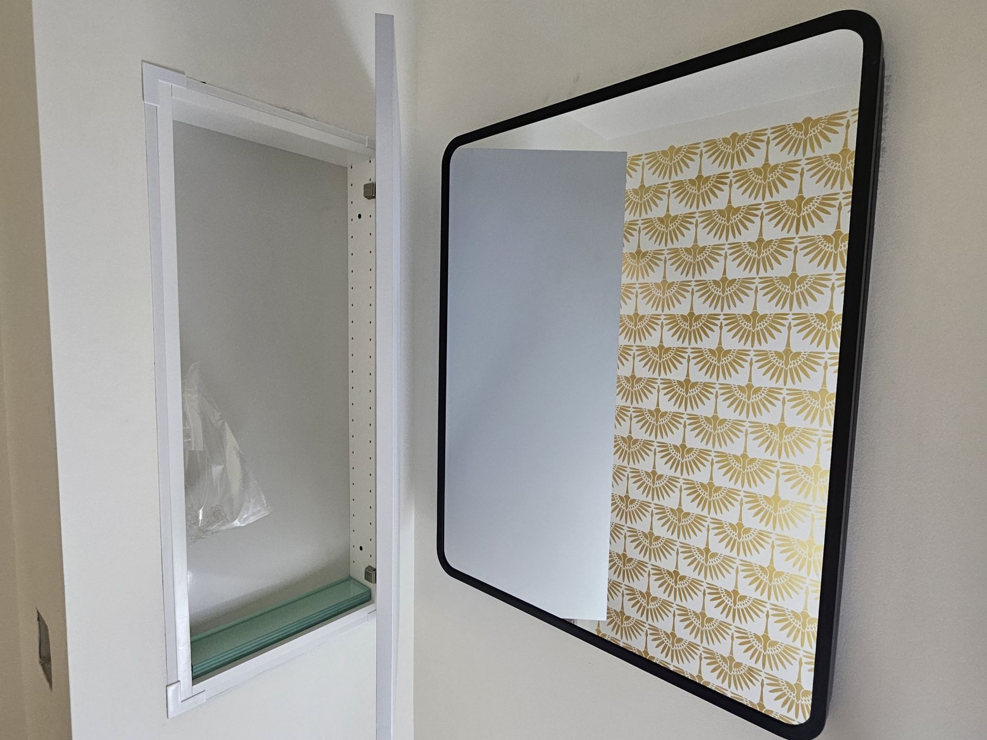 A black-framed mirror next to an open medicine cabinet on a cream-colored wall. The mirror reflects a wallpaper with gold geometric patterns.