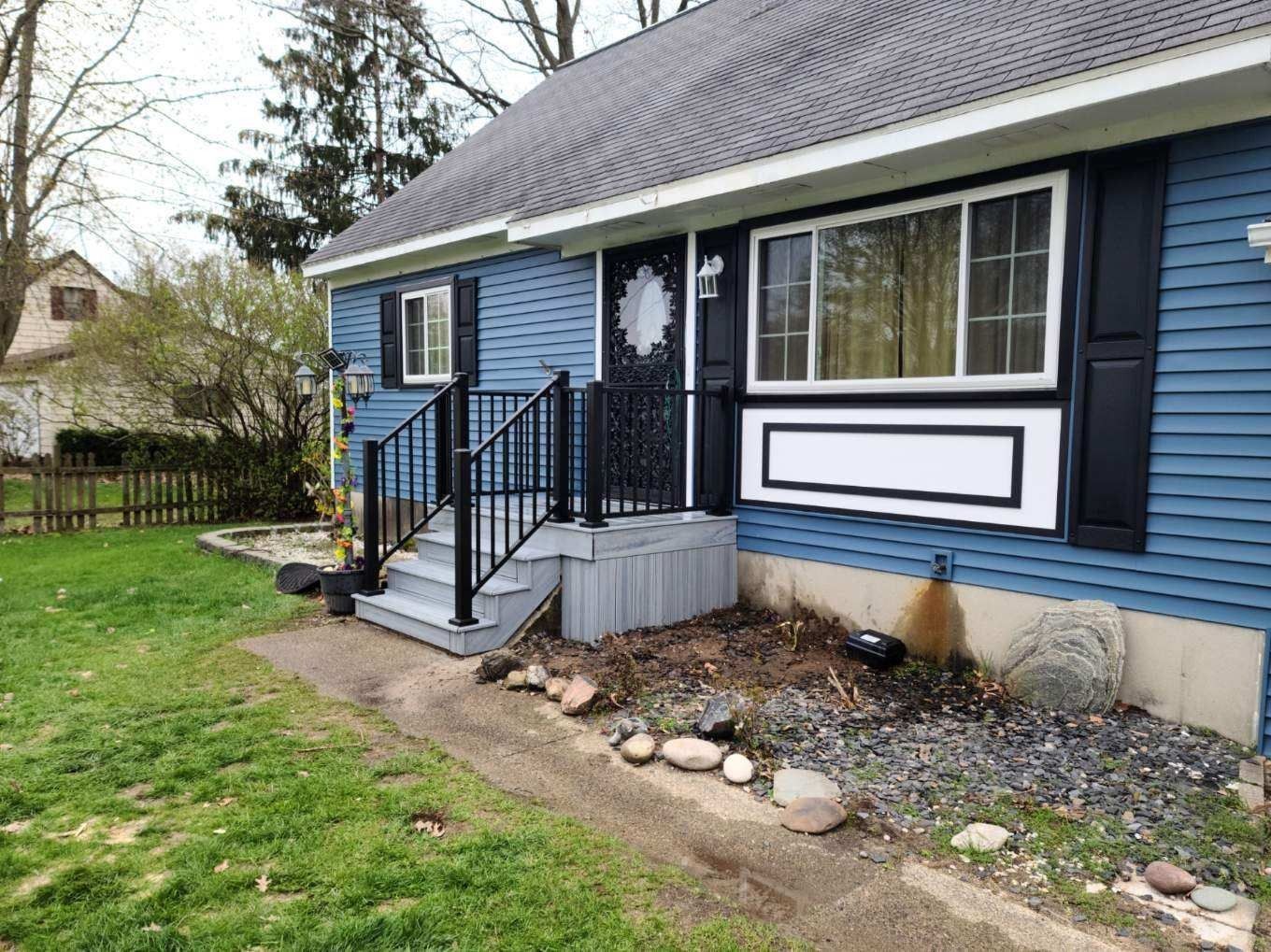 Blue house with black shutters and railing. A front porch and small yard with grass, rocks, and a fence.