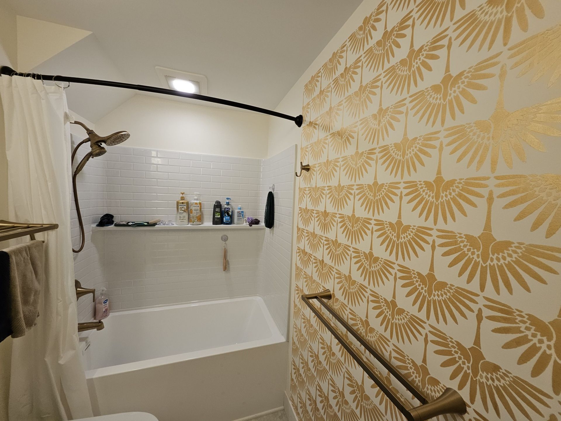 Bathroom with white and gold patterned wallpaper, a bathtub, shower, and bronze fixtures.