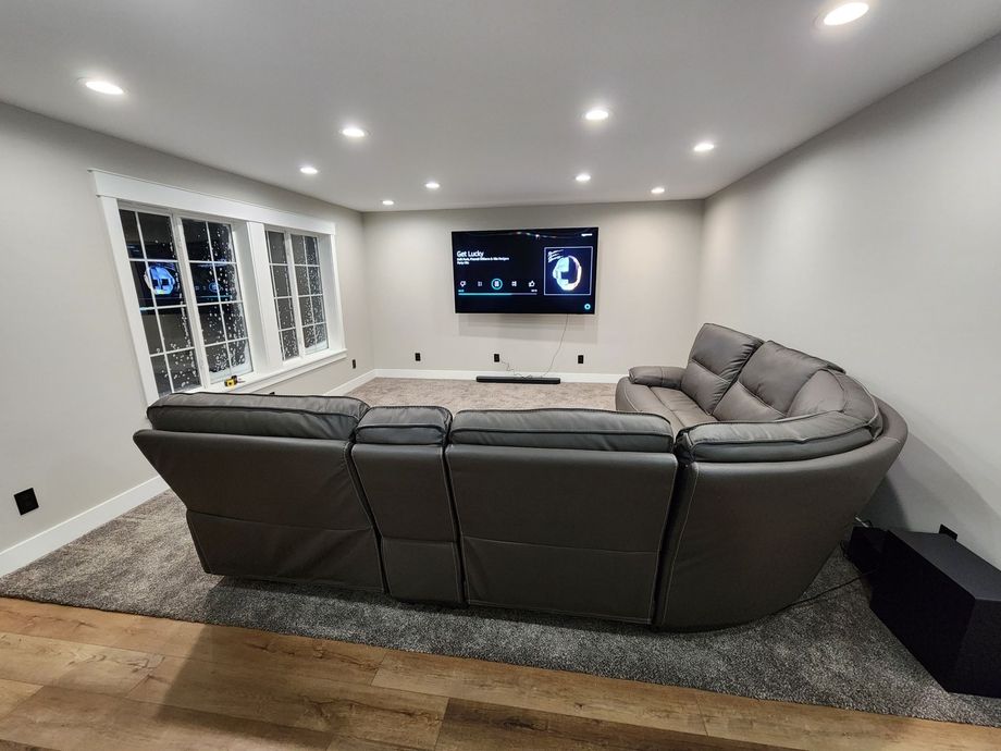 Grey leather sectional sofa facing a large TV mounted on a gray wall in a modern living room with recessed lighting and a large window.