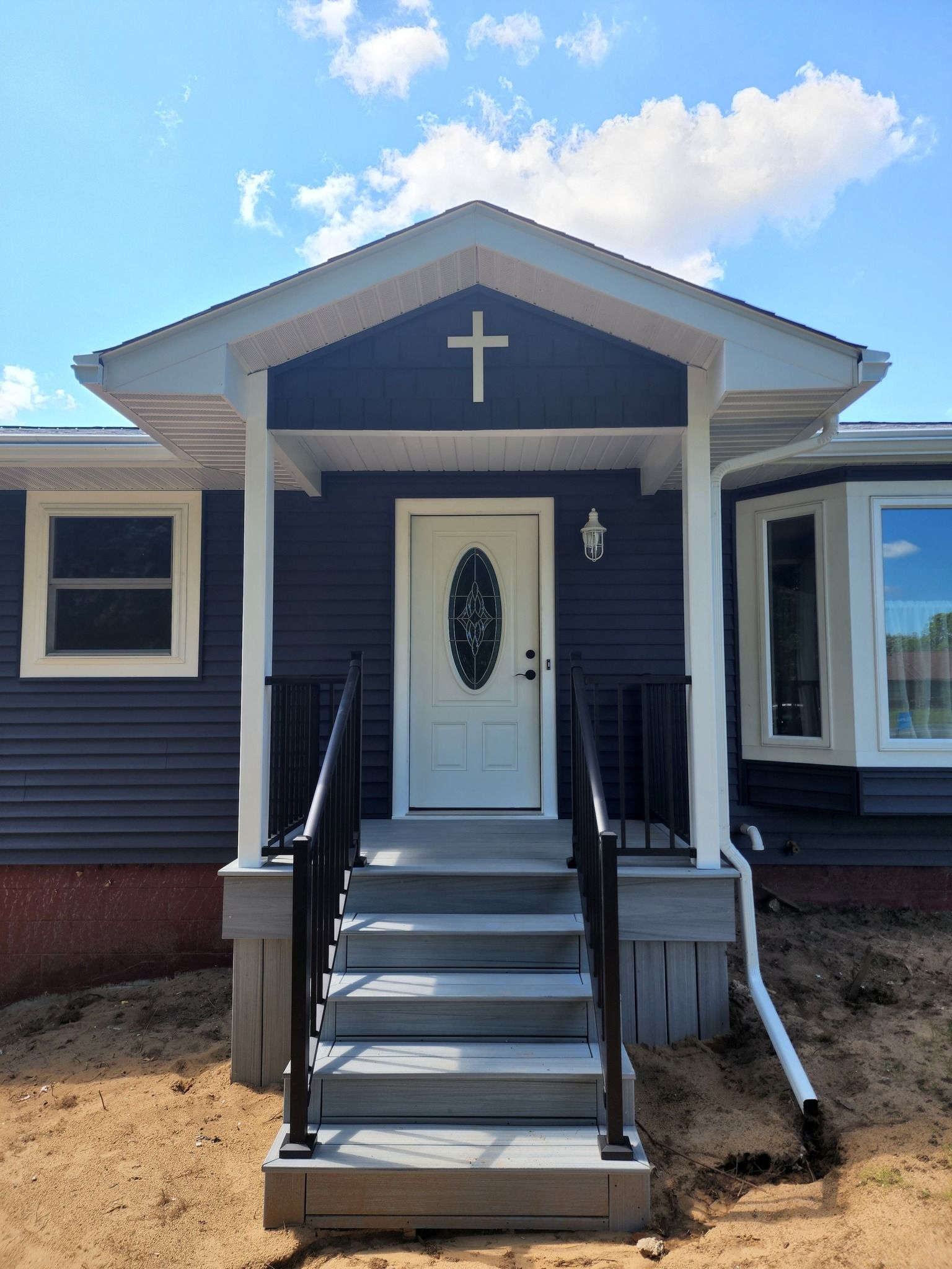 A home with a white door and gray steps, topped with a porch featuring a cross. The house is navy blue with white trim.