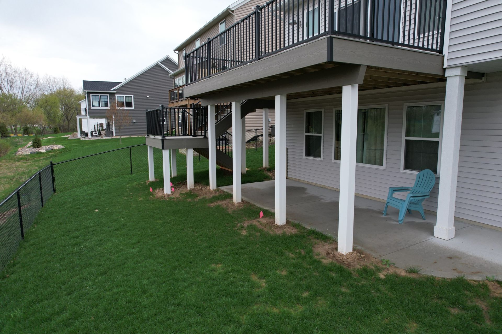 A backyard with green grass, a black fence, and a two-story house with a deck above a concrete patio. A blue chair sits on the patio.