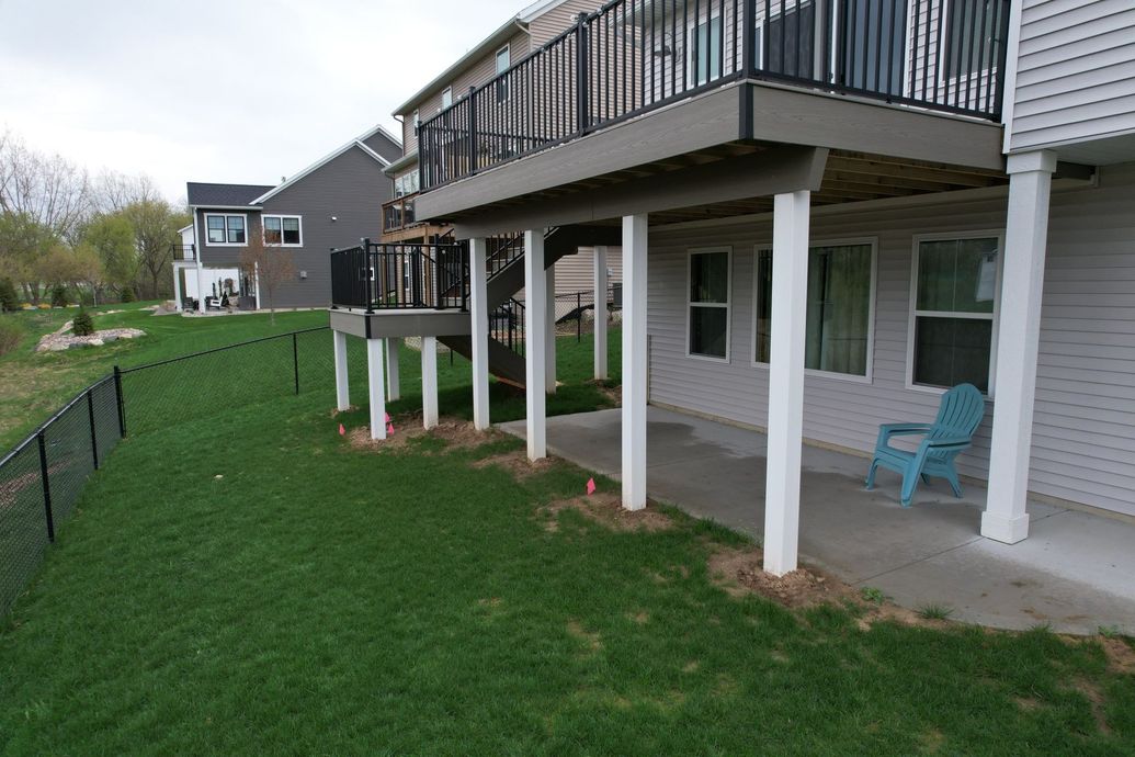 A backyard with a two-story house and a deck. A small fenced-in yard of green grass, with a blue chair near the house.