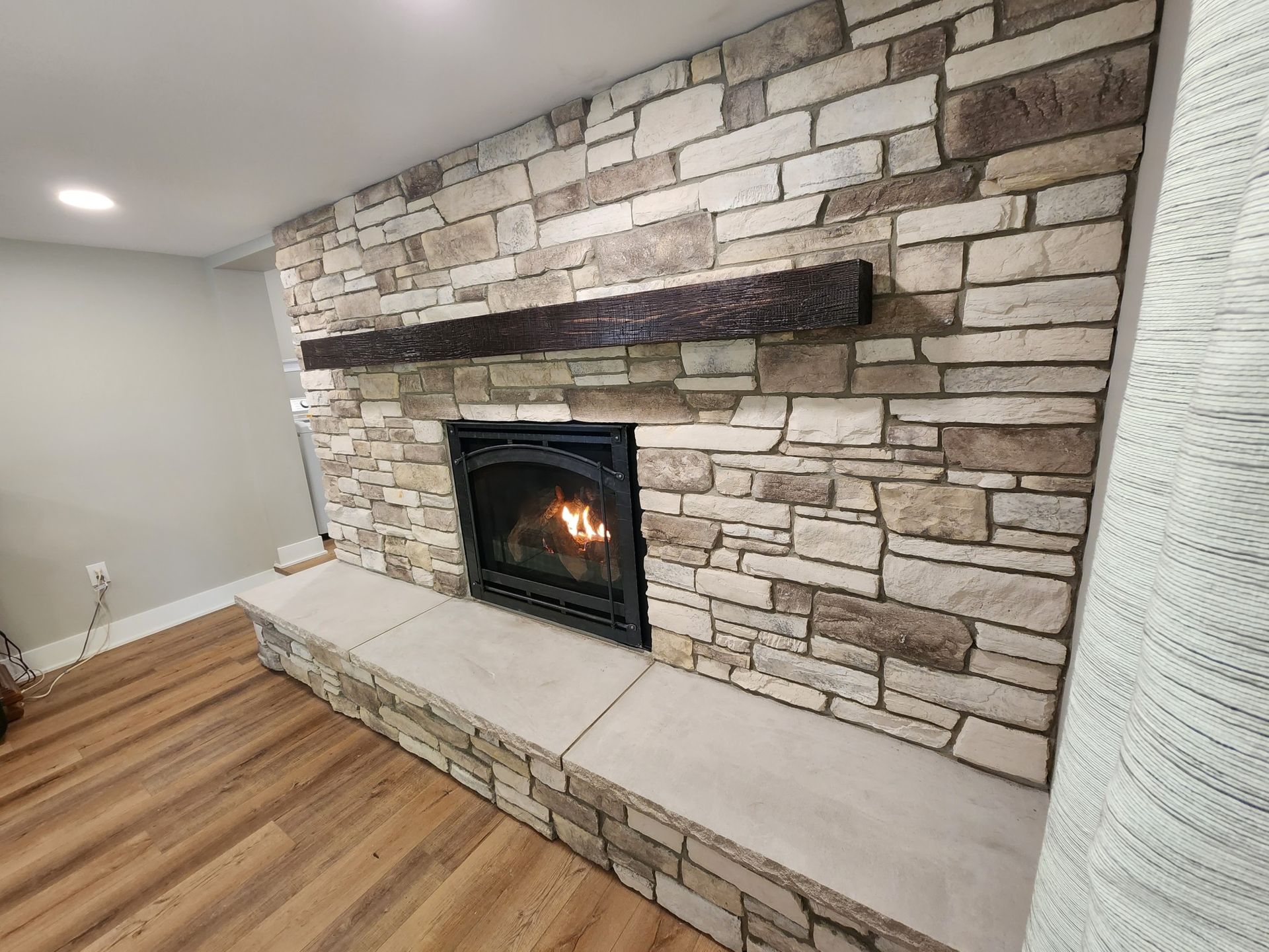 Fireplace with stacked stone surround and a dark wooden mantel. The fire is lit in the black fireplace, and the hearth is light stone.