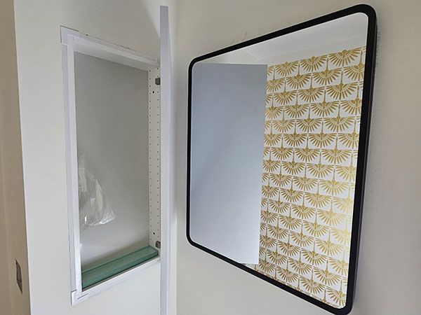 A modern bathroom with a black-framed mirror and an open recessed cabinet, reflecting patterned wallpaper.