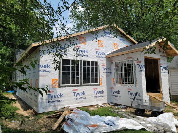 House under construction, wrapped in white Tyvek. Windows installed, with a small porch area being framed. Green trees surround.