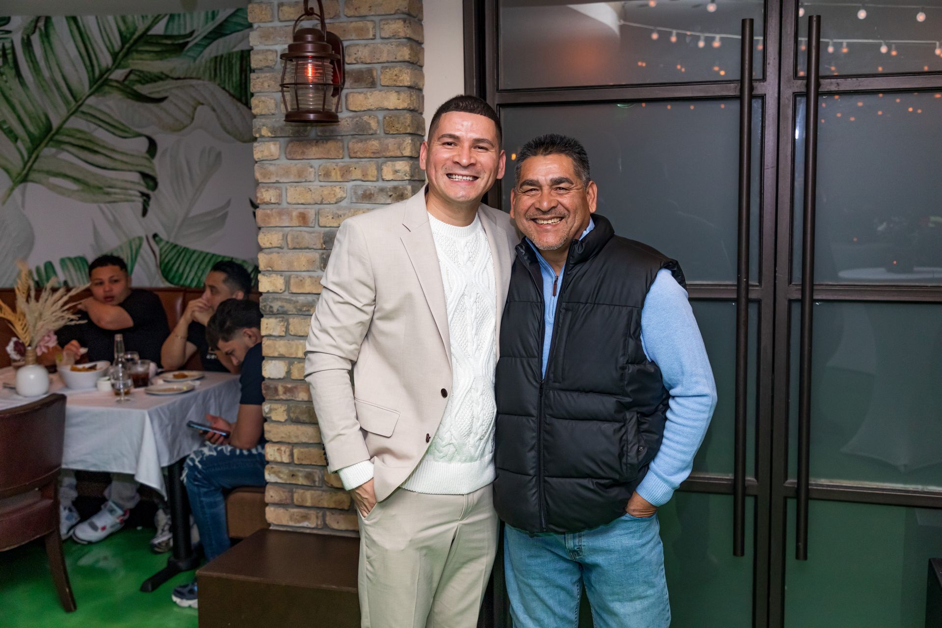 Two men smiling, one in a tan suit, the other in a vest and jeans, inside a restaurant.