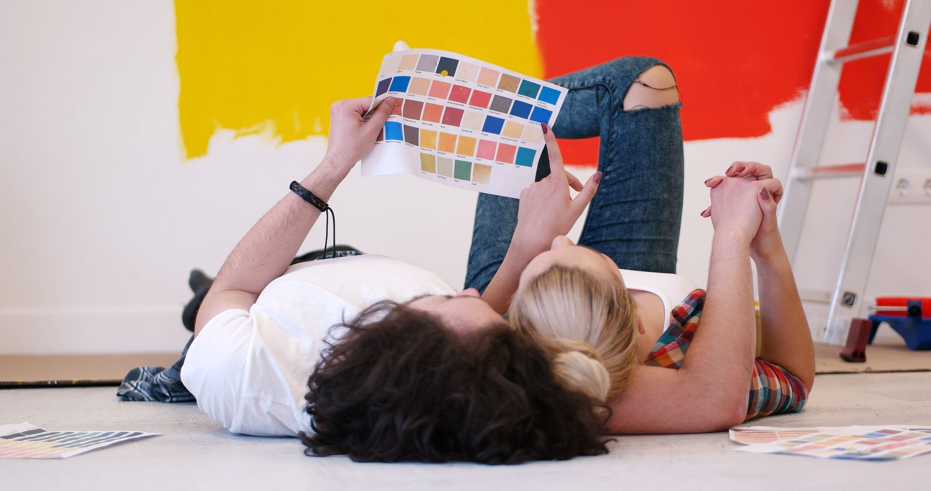 Couple on floor, looking at paint swatches in front of a partially painted wall, ladder in background.