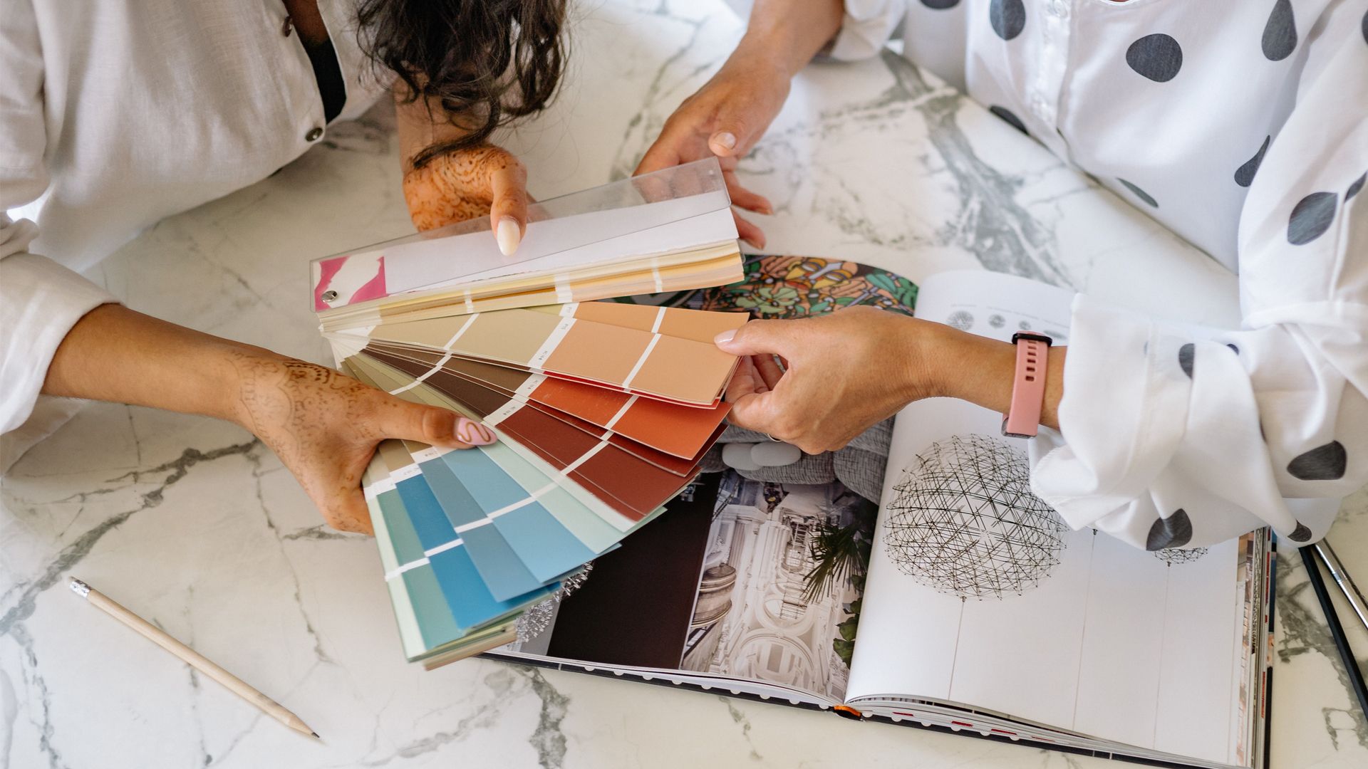 Hands holding paint swatches and a design book on a marble table.