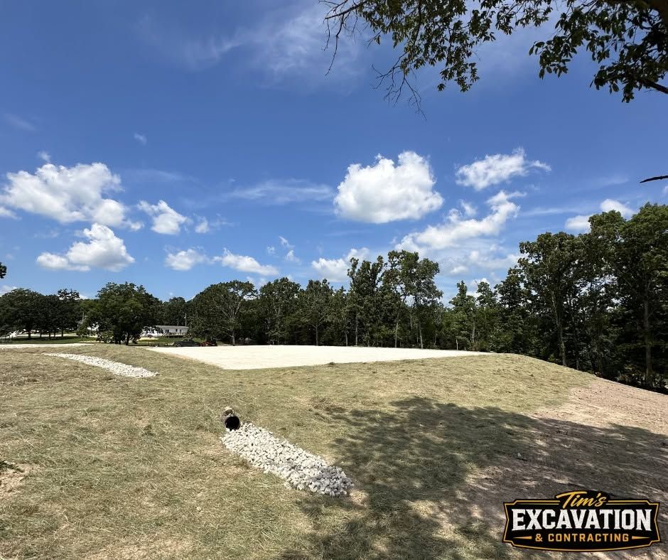Construction site with a leveled dirt field, gravel trench, trees, and blue sky with clouds