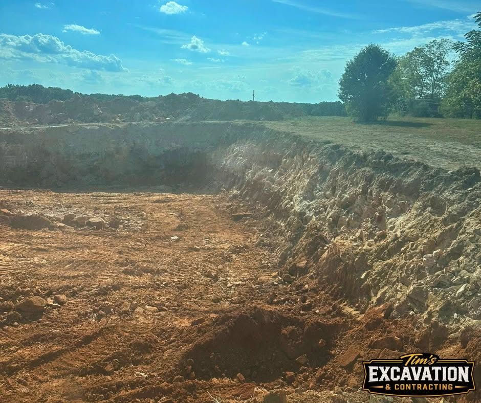 Large dirt excavation pit in a rural field under a blue sky, with an excavation logo in the corner