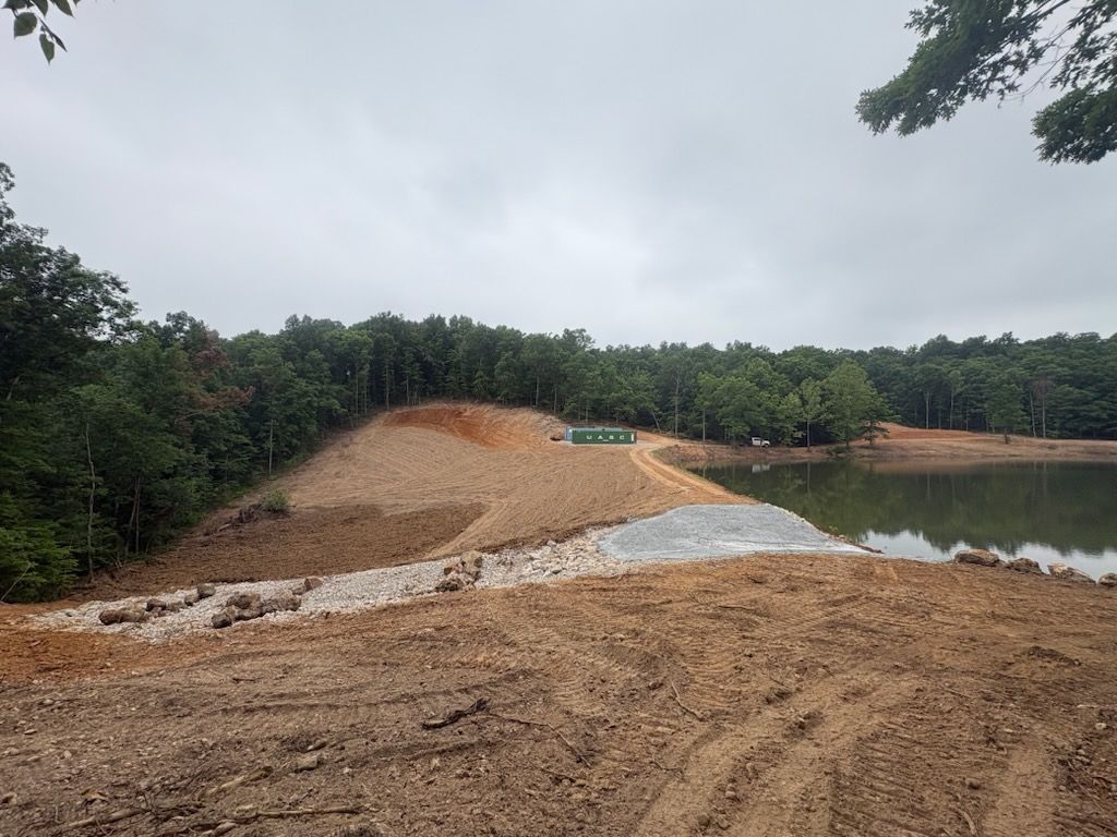 Gravel embankment under construction beside a pond, with forest and cloudy sky in the background