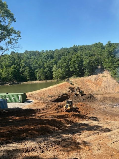 Construction site with excavator on dirt slope beside a green lake and forested hillside