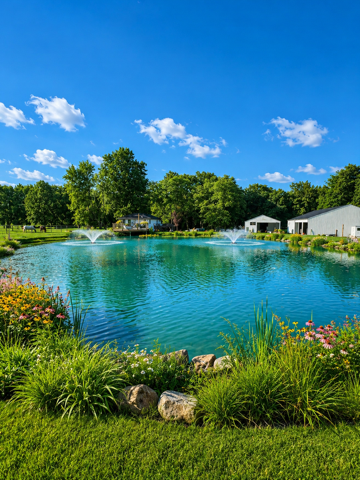 Bright blue pond with fountains, green lawn, flowers, and trees under a sunny sky