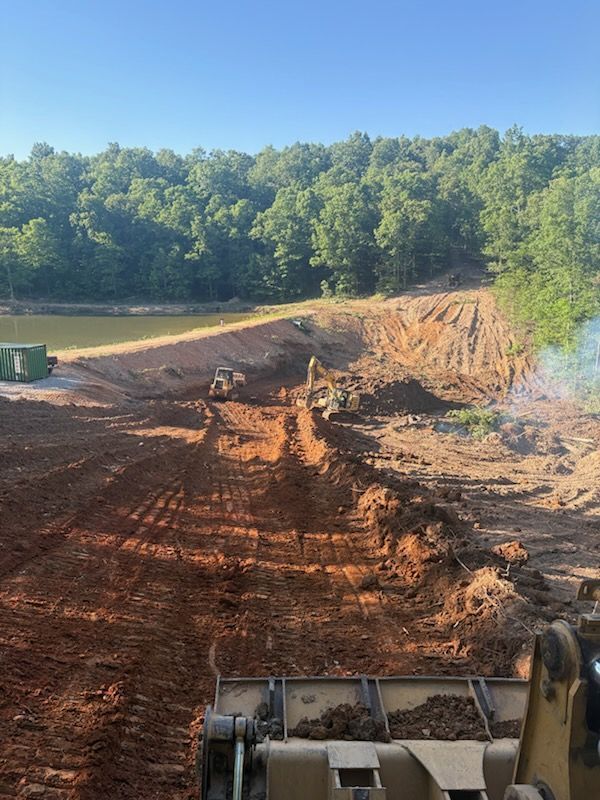 Excavator grading a red dirt path beside a wooded pond under a clear blue sky