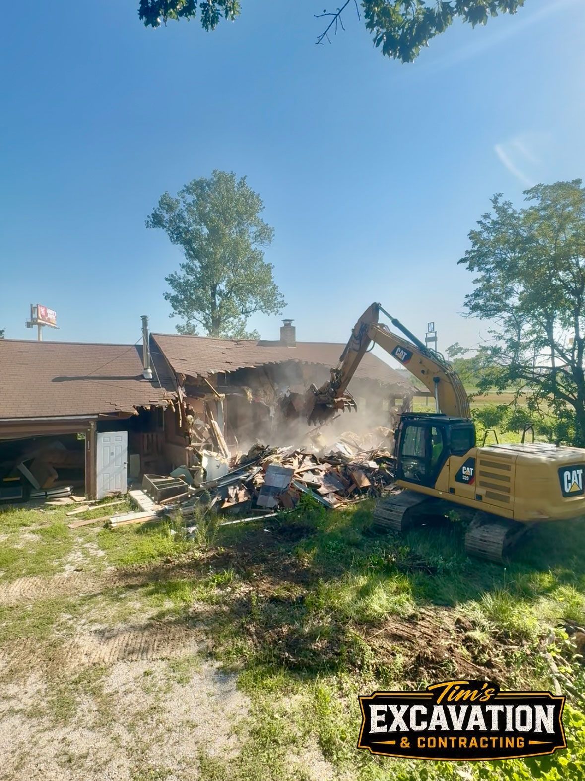 Excavator demolishing a damaged house, with dust and debris flying under a blue sky.