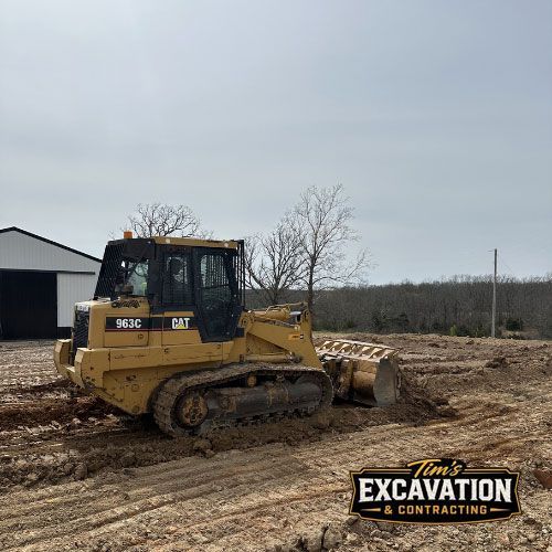 Yellow CAT bulldozer grading muddy dirt lot near a barn under a gray sky