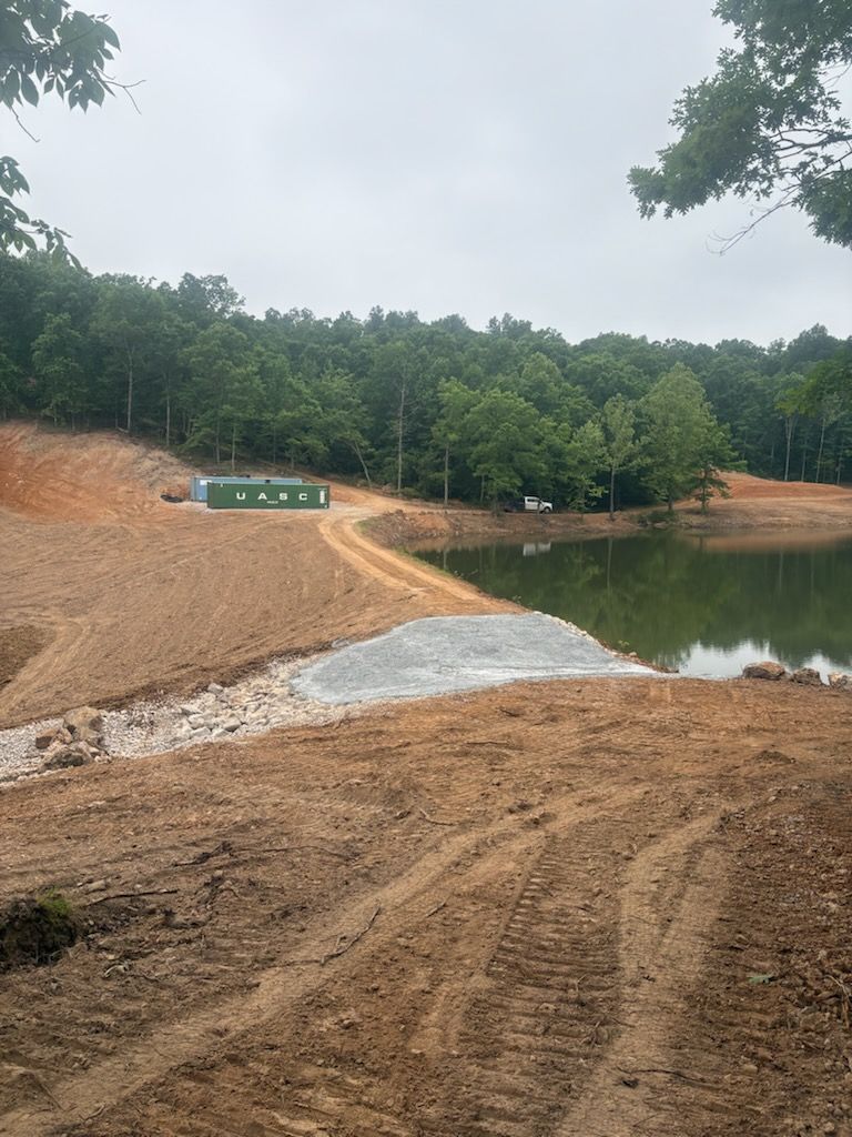 Construction site beside a pond, with dirt embankments, gravel, and a tree-lined hillside under cloudy skies.