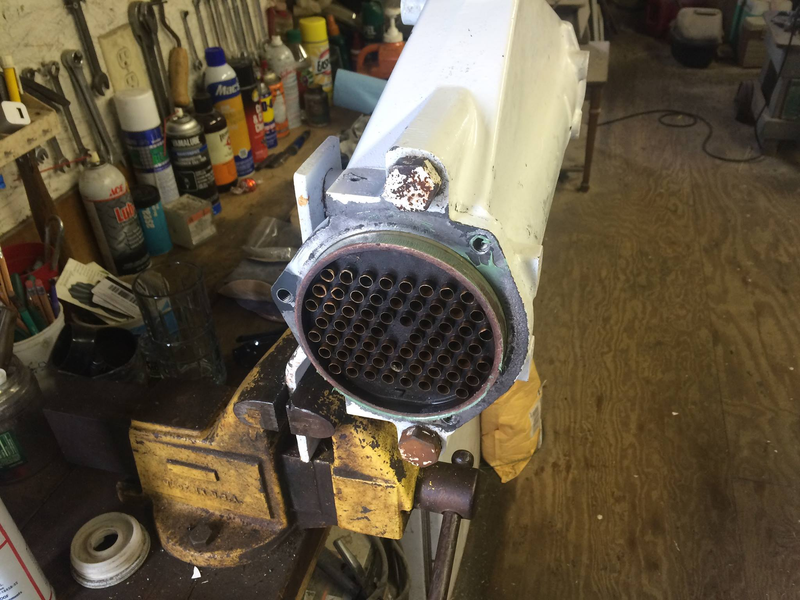 A metal cylindrical heat exchanger core held in a yellow vise on a workbench cluttered with tools and workshop supplies.