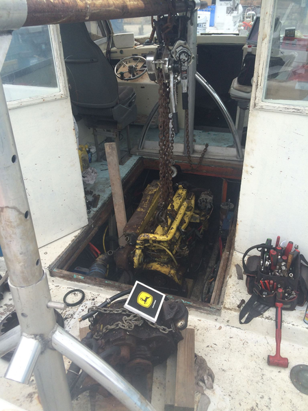 A yellow marine engine being lifted by a chain hoist out of an open floor hatch on a boat deck.