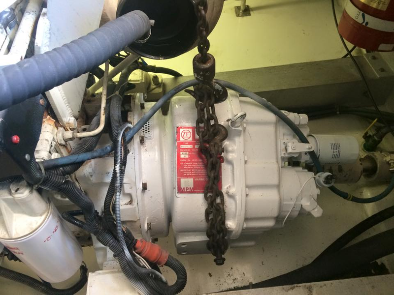 A marine transmission unit with a red manufacturer plate is suspended by a heavy metal chain in an engine compartment.
