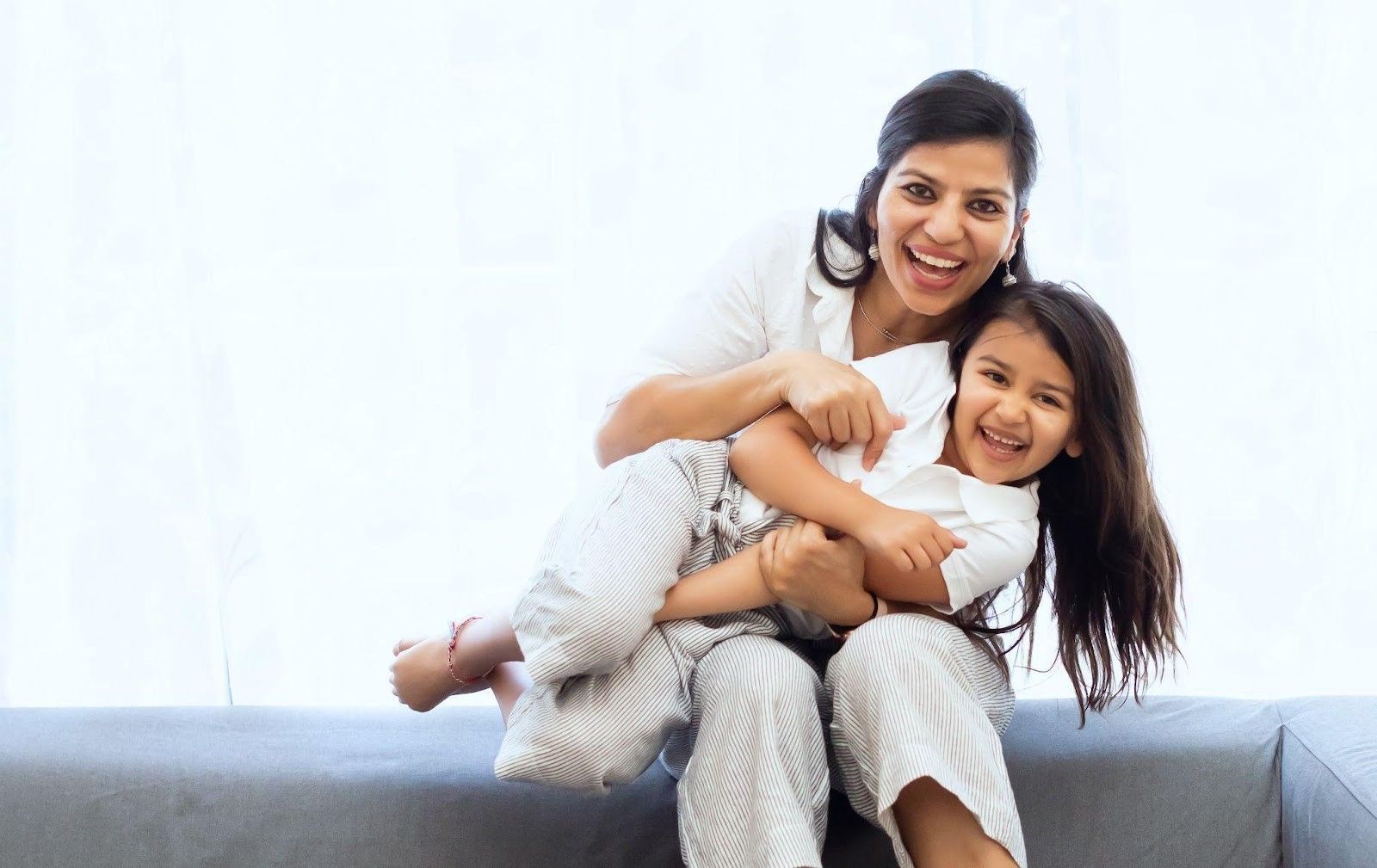 A person sitting on a sofa, laughing while playfully hugging a child in their lap against a bright white background.