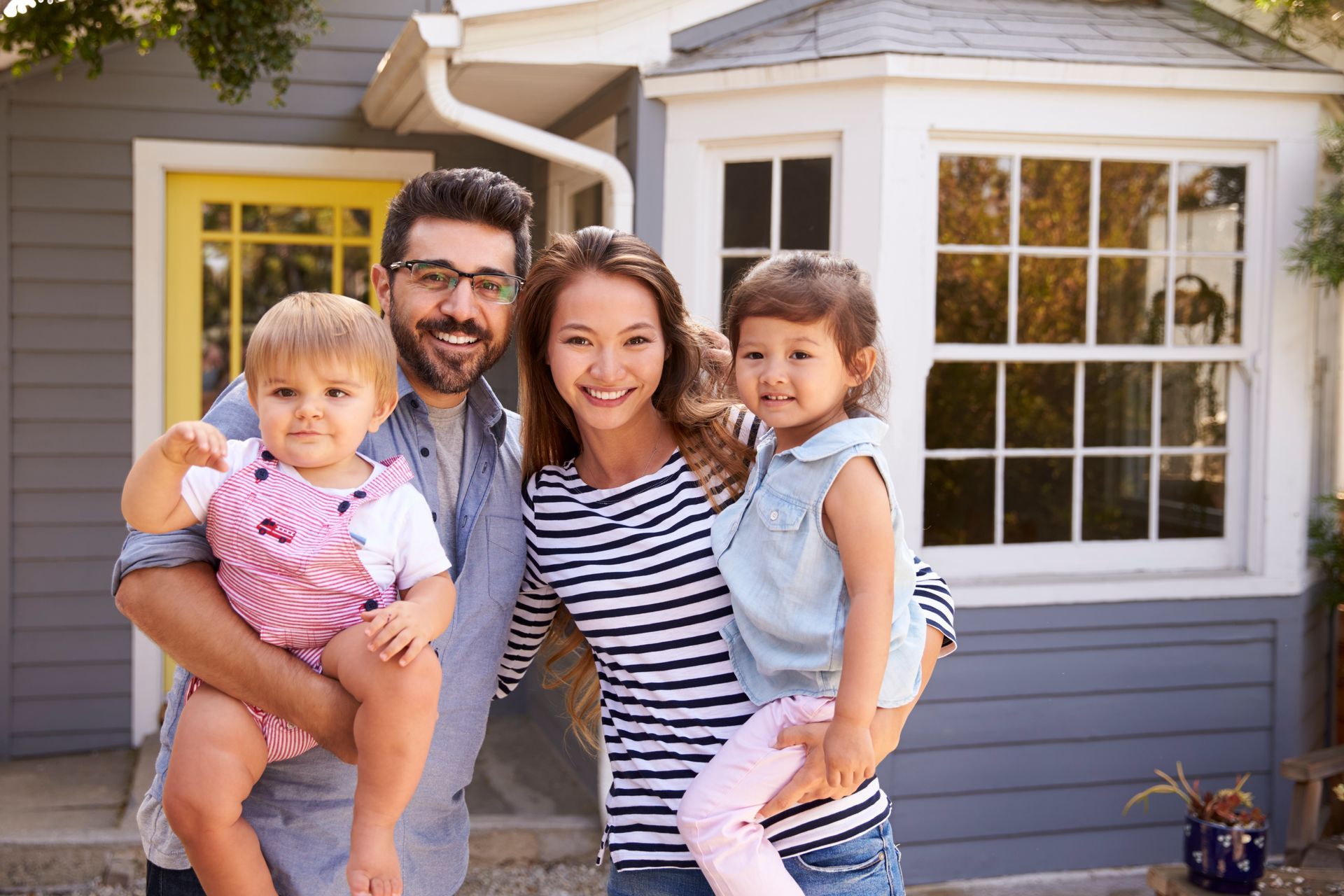 Family of four smiling in front of their home; father holds baby, mother holds child.