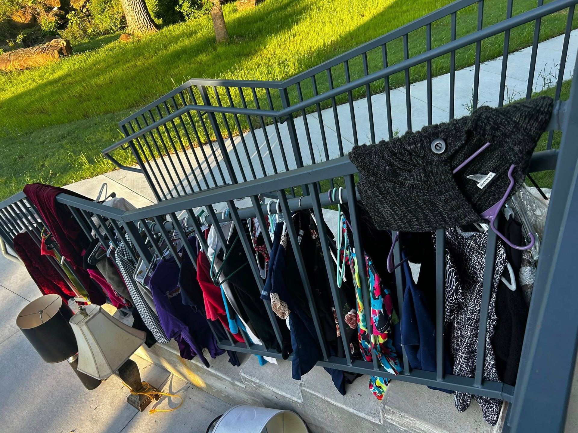 Clothes hanging on a gray metal railing on stairs, suggesting a yard sale. Sunlight.