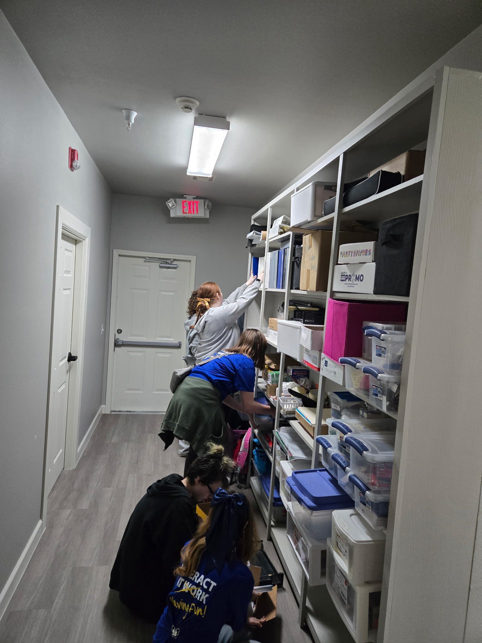 People in a storage room, reaching for items on shelves. White shelving against a gray wall.