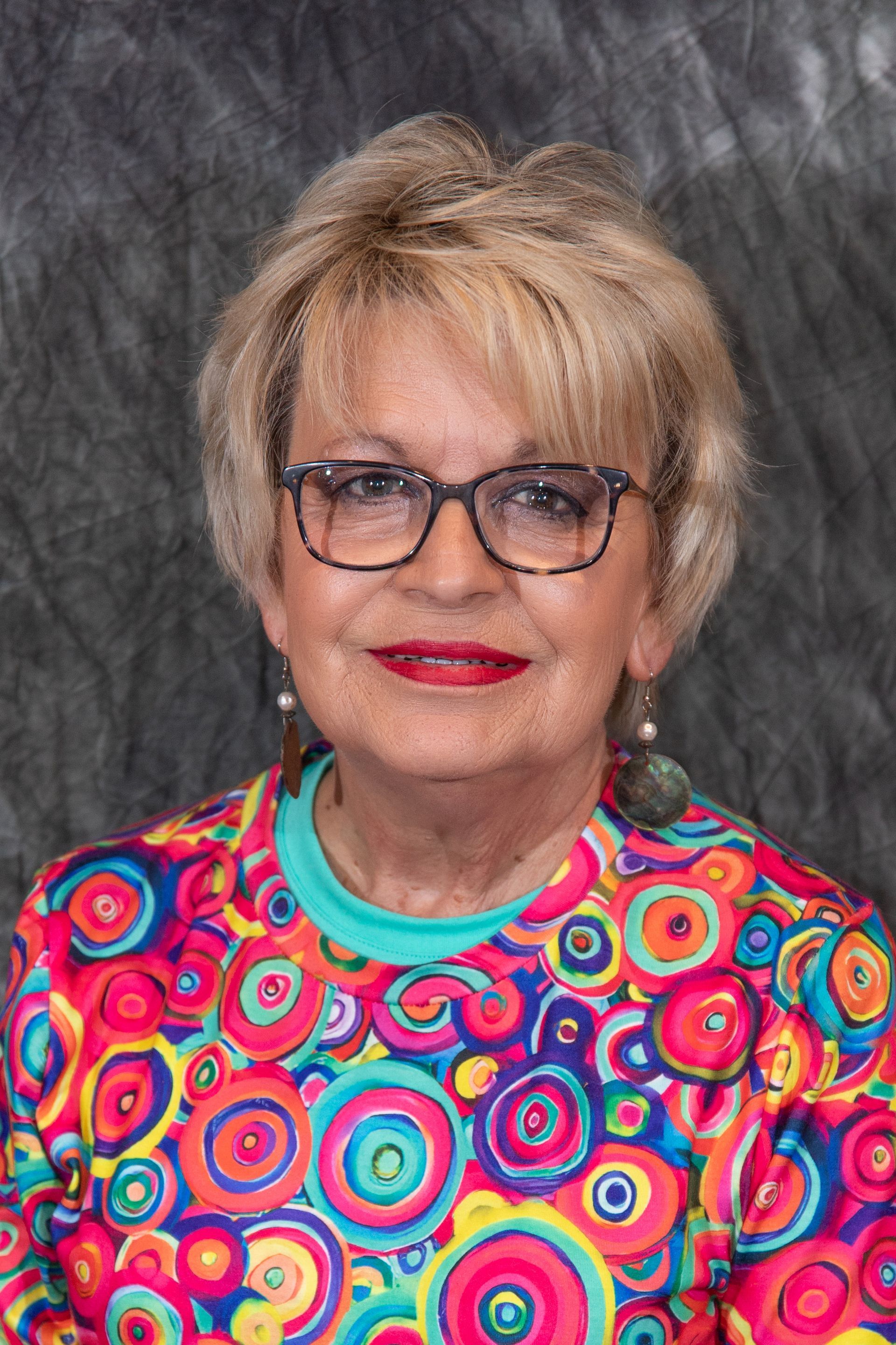 Woman with short, layered brown hair, wearing a patterned top with colorful embroidery, smiles at the camera.
