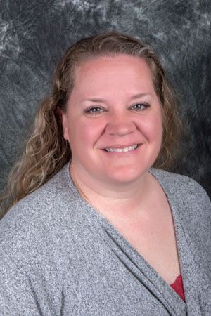 Woman with light skin, smiling, wearing a gray sweater, standing in front of a mottled gray background.