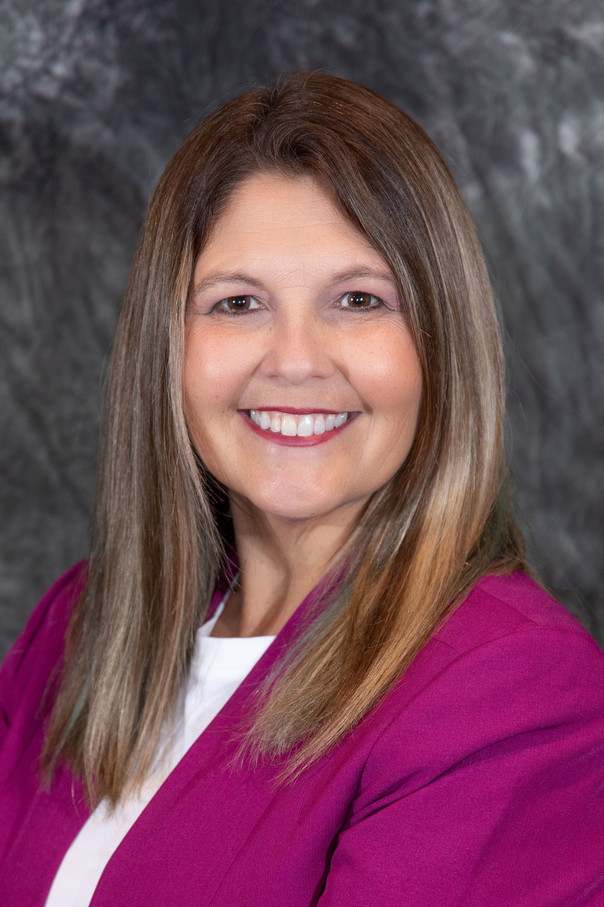 Woman with blonde hair wearing a black blazer over a red top smiles at the camera against a gray backdrop.