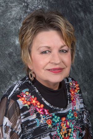 Woman with short, layered brown hair, wearing a patterned top with colorful embroidery, smiles at the camera.
