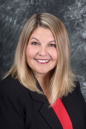 Woman with blonde hair wearing a black blazer over a red top smiles at the camera against a gray backdrop.