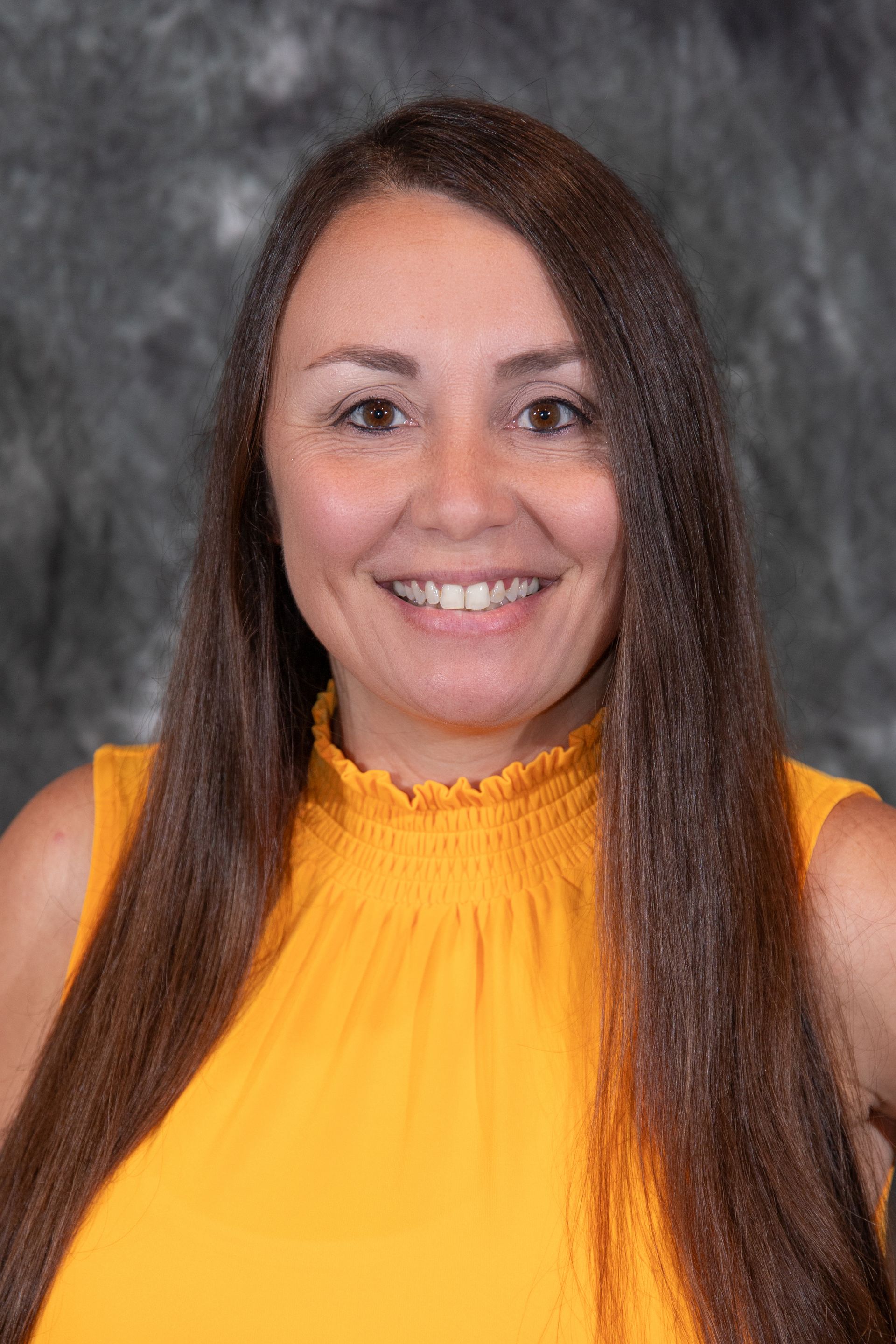 Woman with long brown hair smiling, wearing a red top, against a mottled gray backdrop.