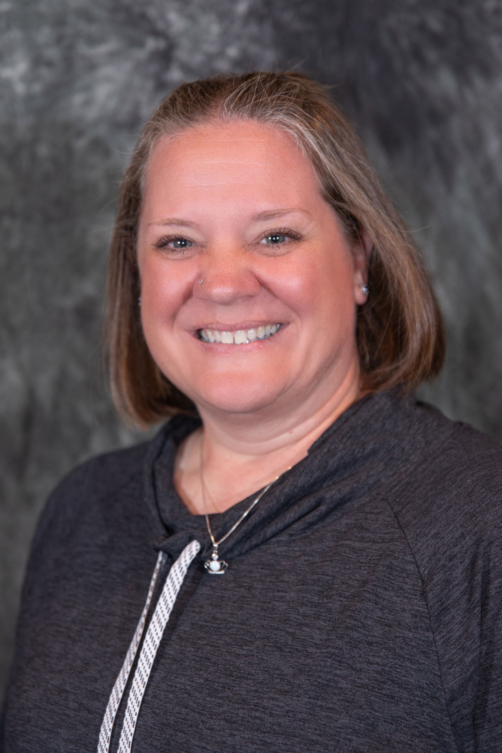 Woman with light skin, smiling, wearing a gray sweater, standing in front of a mottled gray background.