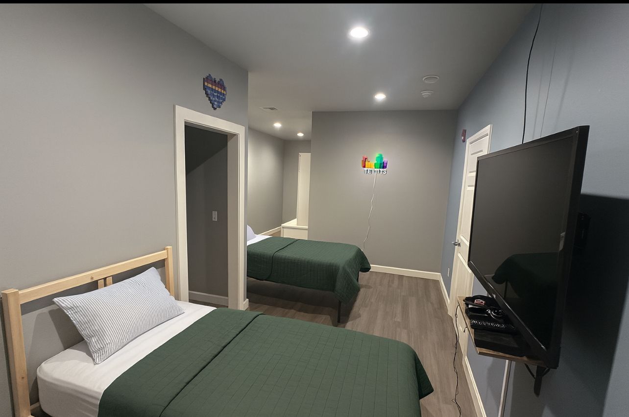 Two beds with green bedding in a gray-walled room, TV on wall, and open doorway.