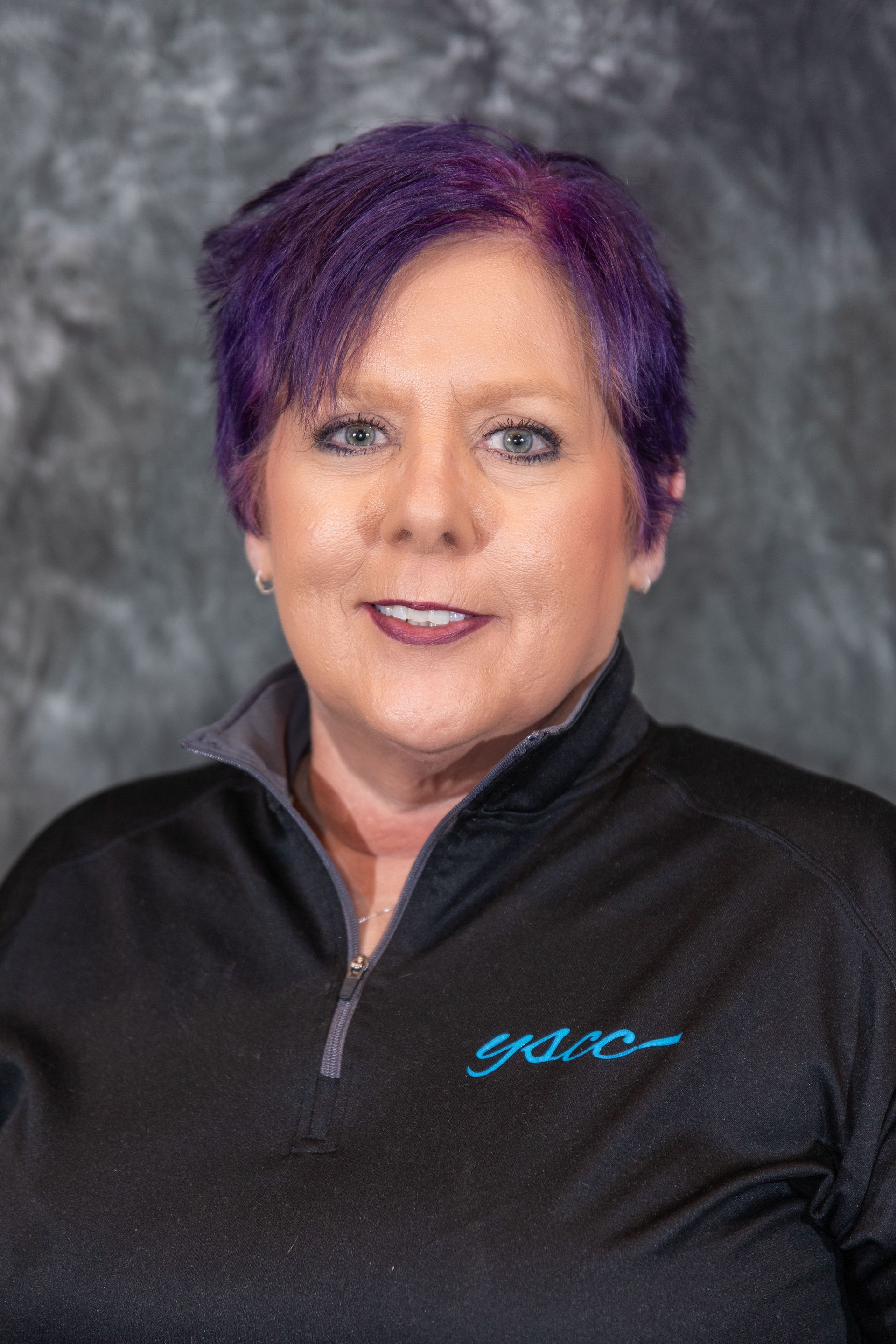 Woman with purple hair smiling, wearing a black shirt with a logo, against a mottled gray backdrop.