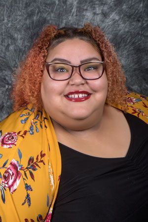 Woman smiling, wearing glasses and a floral yellow jacket over a black top, against a mottled gray backdrop.